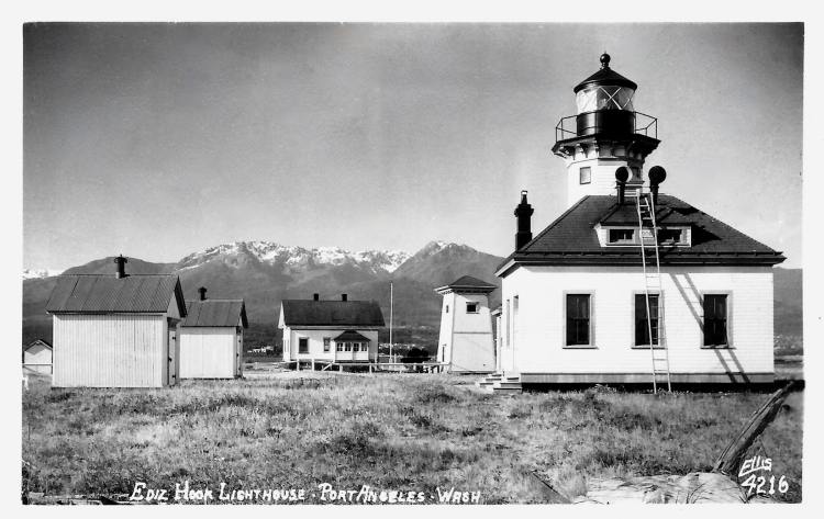 Ediz Hook Lighthouse in Port Angeles, Washington, with the lighthouse tower and several surrounding buildings, set against a backdrop of snowy mountains.